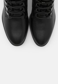 Bottes en cuir noir à finition lisse, avec un laçage distinctif et des bouts arrondis, sur fond blanc.