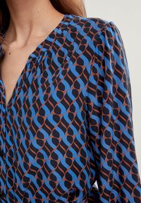 Kuosi blouse dengan desain bergelombang berwarna biru dan coklat di latar belakang hitam. Bahan ringan dengan sedikit kilau. Memiliki leher V.