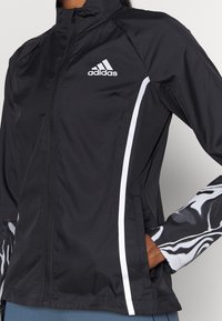 Musta adidas-takki, jossa vetoketjulla suljettava etuosa, valkoiset korostukset sivuilla sekä hihoissa mustavalkoinen marmoroitu kuvio.