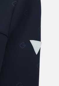 Mornarska tkanina z embosiranimi vzorci logotipa; na roka je bela trikotna nalepka logotipa Guess; gladka tekstura, prilegajoča zasnova.