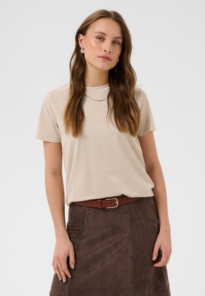 Femme aux longs cheveux bruns portant un haut beige à manches courtes, une jupe en daim marron avec ceinture, un collier argenté et des boucles d'oreilles à anneaux sur un fond uni.