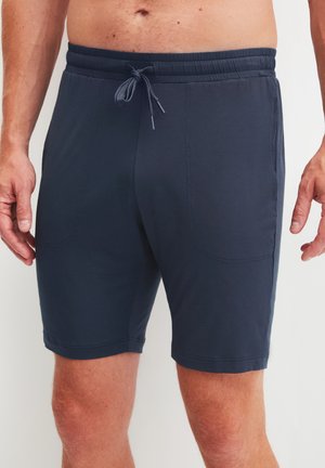 Uomo che indossa pantaloncini blu navy con coulisse e tasche a patch frontali, in piedi con le braccia rilassate ai lati.