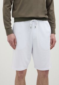 Shorts bianchi in cotone con vita elastica e cordoncino, dotati di punte metalliche. Indossati con una maglietta a maniche lunghe verde oliva.