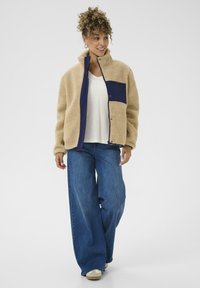 Veste sherpa beige avec poche navy, fermeture à pression et col haut ; associée à un jean bleu à jambes larges et une chemise blanche.