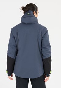 Blå och svart isolerad jacka med hög krage, huva och grön accent på baksidan. Har långa ärmar och en lös passform.