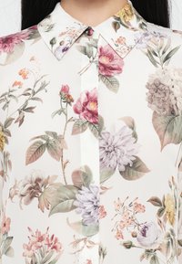 Gros plan d'une chemise blanche avec un imprimé floral comprenant des fleurs roses, violettes, jaunes et beiges ainsi que des feuilles vertes, avec un col pointu.