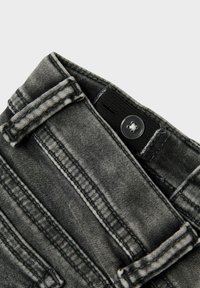 Grå denimjeans med en strukturerad yta, med knappstängning och bältesloopar, som framhäver kontrasterande sömmar längs kanterna.