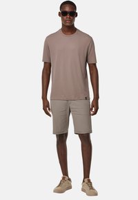 T-shirt a maniche corte di cotone marrone chiaro abbinata a pantaloni corti beige. Il modello indossa occhiali da sole e sneaker beige chiare. Un outfit semplice e casual.