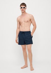Námornícke modré plavky s elastickým pásom, detailom na stiahnutie, bočnými vreckami a malým logom. Nosí ich model bez trička.