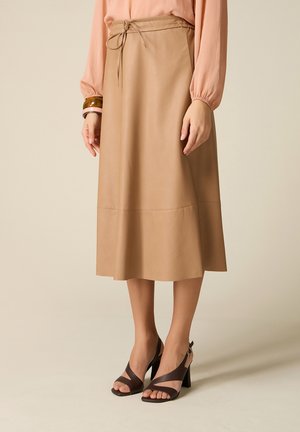 Donna che indossa una gonna midi beige con cintura annodata in vita, una camicetta color pesca a maniche lunghe, sandali con tacco marroni e due braccialetti al polso sinistro.