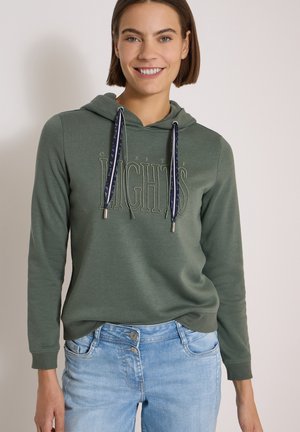 Femme souriante portant un sweat à capuche vert avec des cordons et le texte "Lights", associée à un jean bleu clair sur un fond uni.