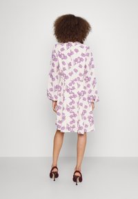 Weißes Kleid mit lila Blumenmuster, langen Ärmeln und einem fließenden Design. Kombiniert mit burgunderfarbenen Absätzen mit gewebter Textur.