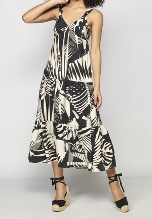 Femme portant une robe maxi sans manches noir et blanc avec des motifs abstraits de feuilles et des sandales compensées espadrilles noires avec liens à la cheville.
