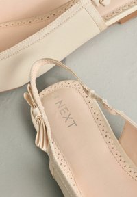 Chaussures plates beiges en matériau synthétique avec un bord festonné, un nœud décoratif et une semelle intérieure rembourrée de couleur claire portant le nom de la marque "NEXT".