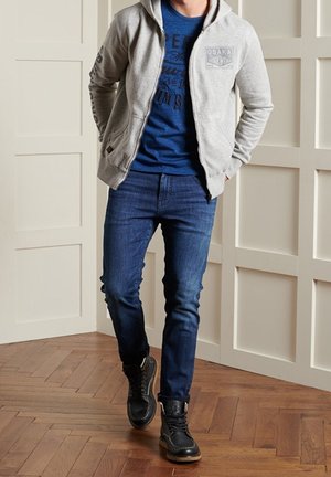 Hombre con sudadera con capucha gris con cremallera, camiseta azul con diseño gráfico, vaqueros azul oscuro y zapatillas negras de caña alta de pie sobre suelo de madera.