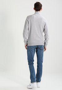 Grijze sweatshirt met geribde manchetten en zoom, gecombineerd met blauwe jeans en witte sneakers, van achteren weergegeven tegen een neutrale achtergrond.