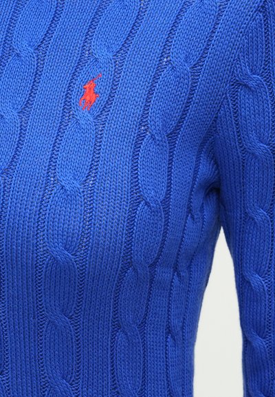 Kék kötött pulóver, amely fonott mintával és egy kis piros logóval díszített a bal mellrészen. A közelkép kiemeli a textúrát és a varrást.