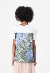 T-shirt à manches courtes en blanc avec un motif patchwork géométrique présentant des motifs verts, bleus et noirs au dos. En matière de coton.