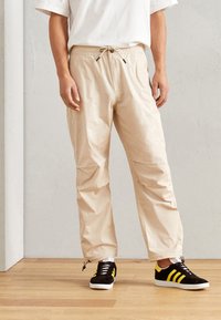 Beige snøretrækkebukser lavet af letvægtsstof, med sidelommer og manchetter, parret med sorte og gule sneakers.