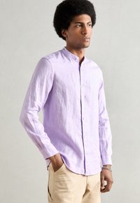 Man met afro-haarstijl die een lichtpaarse mandarin-kraagshirt en beige broek draagt, poseert met één hand in de zak.