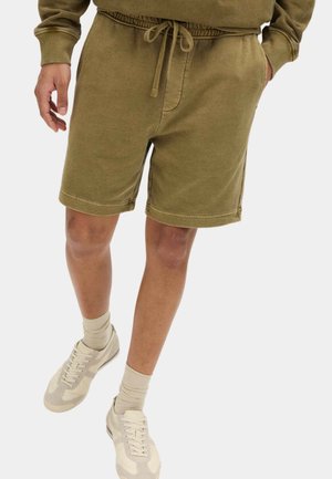 Uomo che indossa pantaloncini verdi oliva con coulisse, calzini beige e sneakers beige, con una mano in tasca su sfondo bianco.
