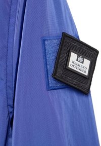 Ärmel einer blauen Nylonjacke mit einem rechteckigen, strukturierten schwarzen Patch, auf dem ein weißes Etikett mit der Aufschrift "WEEKEND OFFENDER" zu sehen ist.