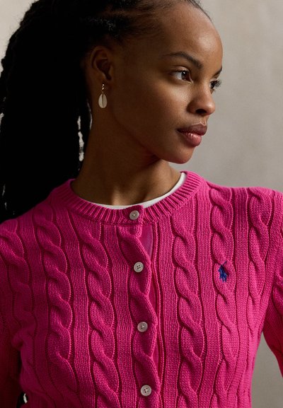 Ροζ ζακέτα πλεκτή με στρογγυλή λαιμόκοψη, πέντε λευκά κουμπιά και μικρό μπλε λογότυπο στην αριστερή πλευρά. Ομαλή υφή σε όλο το ύφασμα.