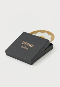Caja negra de Versace Rosenthal parcialmente abierta que revela el borde blanco y dorado ornamentado de un plato en el interior sobre un fondo blanco.
