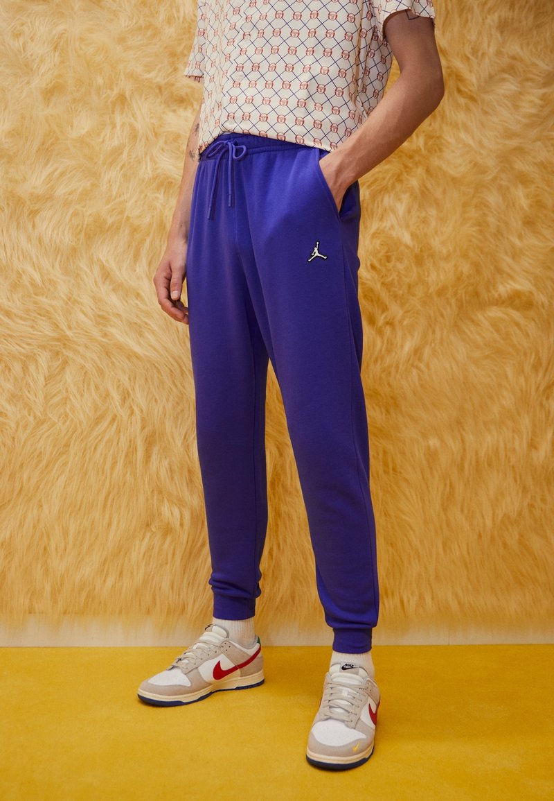 Lila joggers med dragsko i midjan, med en svart Jordan-logotyp. Kombinerat med en mönstrad t-shirt med kort ärm och beige Nike-sneakers.