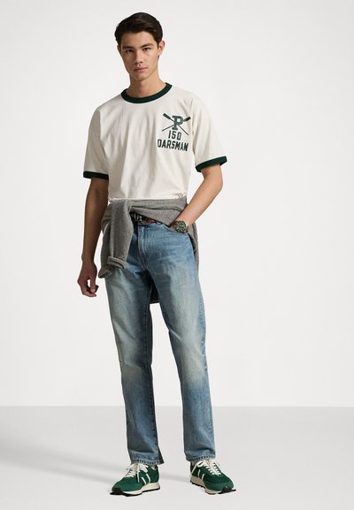 T-shirt blanche avec col vert et texte, jeans bleu clair, et baskets vertes. Pull gris noué autour de la taille. Design simple et décontracté.