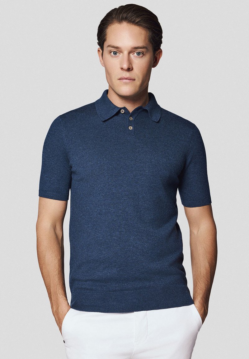 Granatowa koszulka polo z krótkim rękawem, subtelnej teksturze, z trzema guzikami i klasycznym kołnierzykiem, zestawiona z białymi spodniami.