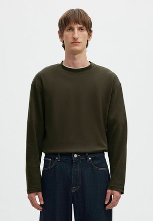 Joven con cabello corto y castaño que lleva una camisa de manga larga verde oscuro y vaqueros azul oscuro, de pie frente a un fondo liso.