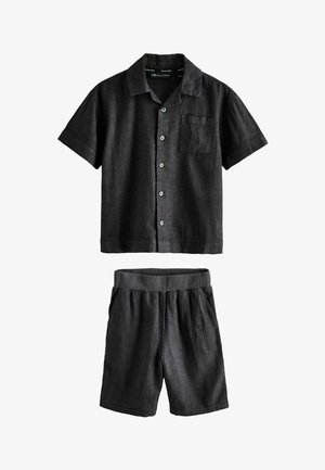 Dospievajúci chlapec stojaci v čiernej košeli s krátkym rukávom na gombíky, zodpovedajúcich šortkách a hnedých sandáloch so sponou, pozerá sa zo strany s rukami v kapsách.