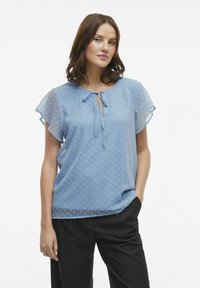 Blusa azzurro chiaro con un motivo a pois testurizzato, maniche corte a sbuffo e un dettaglio a nodo sul collo. Indossata con pantaloni neri.