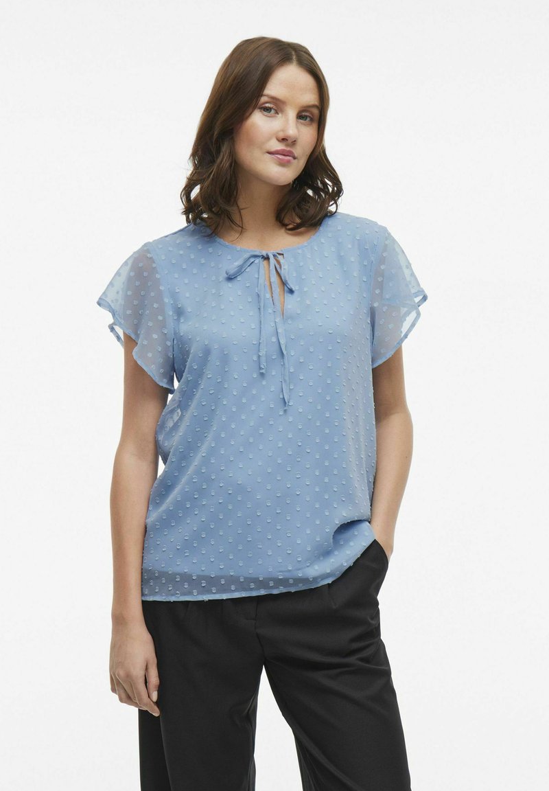 Blusa azzurro chiaro con un motivo a pois testurizzato, maniche corte a sbuffo e un dettaglio a nodo sul collo. Indossata con pantaloni neri.