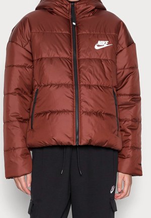 Homme portant une doudoune marron Nike avec une fermeture éclair noire et des poches, associée à un pantalon de jogging noir Nike avec des cordons.