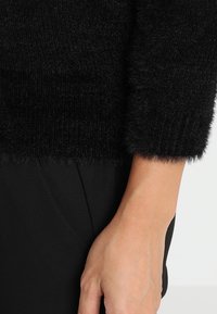 Pull noir en peluche avec une texture côtelée et des manches trois-quarts, associé à un tissu noir lisse. Main reposant contre le tissu.
