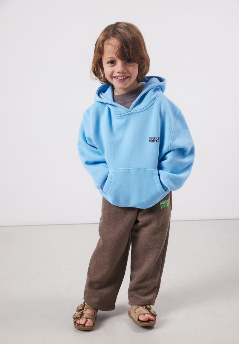 Naeratav laps, kandes helesinist kapuutsi, pruunid püksid logoga ja sandaale, seisab käed taskutes tavalise tausta ees.