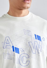 Světlebarevné bavlněné tričko s geometrickým modrým grafickým designem a písmeny "A," "C" a "W." Styl s kulatým výstřihem, jemná textura.