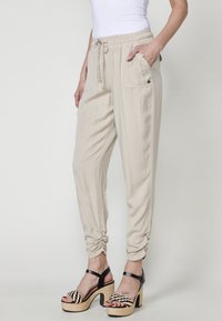 Beige jogger-housut, joissa on kuminauhavärityö, sivutaskut ja kerätyt hihnat. Yhdistetty mustiin korkokenkiin, joissa on kudottuja yksityiskohtia.
