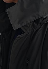 Sort vandtæt jakke med høj krave, lynlås lukkemekanisme, forlommer og glat tekstur. Har en skjult inderlomme.