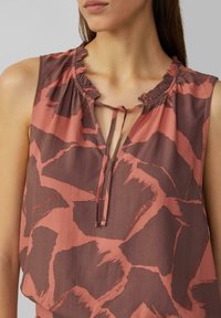 Blouse sans manches, encolure en V, de couleur rouille et marron, avec un col froncé, fermeture par lien et motif abstrait. Tissu léger avec une texture douce.