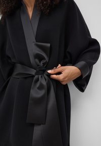 Robe nera con un nastro in satin legato in vita. Il tessuto ha una finitura testurizzata con maniche larghe e una profonda scollatura a V.