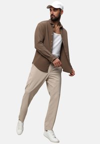 Camicia button-up marrone a costine sopra una canotta bianca, abbinata a pantaloni beige e sneakers bianche. La camicia ha maniche lunghe e colletto ripiegato.