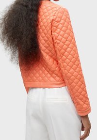 Vadderad orange jacka med dekorativa guldnitar, ribbade manschetter och en kort design. Slät textur med en figursydd silhuett.