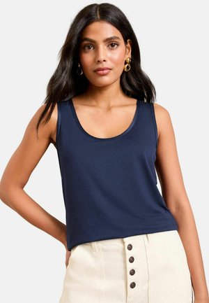 Sieviete ar gariem tumšiem matiem, valkā zelta riņķveida auskarus, tumši zilu piedurkņu neesošu topu un augsta vidukļa krēmkrāsas bikses ar pogām priekšpusē.