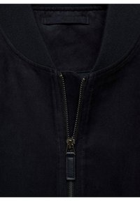 Nahaufnahme einer schwarzen Jacke mit geripptem Kragen und teilweise geschlossenem Metallreißverschluss, der einen schwarzen Leder-Zipper hat.