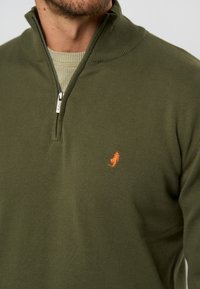 Sudadera de cremallera verde oliva con cuello acanalado, que presenta un pequeño logotipo naranja en el lado izquierdo del pecho y una tela texturizada.