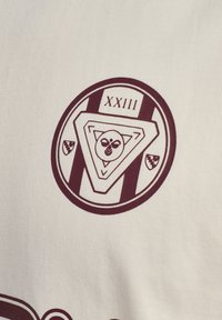Det hvide stof har et rundt vinrødt emblem med et trekantet design, "XXIII" og et billede i midten. Den strukturerede vævning er synlig.