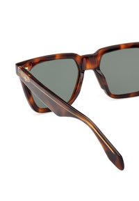 Sluneční brýle ve tvaru želvoviny s geometrickým vzorem, šedými čočkami a elegantním rámem. Kovový detail s logem na spánku.